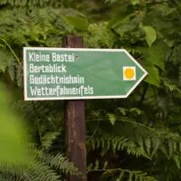 Ein grünes, pfeilförmiges Schild im Wald zeigt nach rechts und trägt die deutschen Aufschriften: „Kleine Bastei, Bertablick, Gedächtnishain, Wetterfahnenfels.“ Das Schild ist von Farnen und grünen Blättern umgeben.