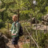 Eine Person mit einem Rucksack, die ein grünes Karohemd und Shorts trägt, lächelt, während sie auf einem felsigen Waldweg mit Metallgeländern wandert, umgeben von Bäumen und viel Grün.