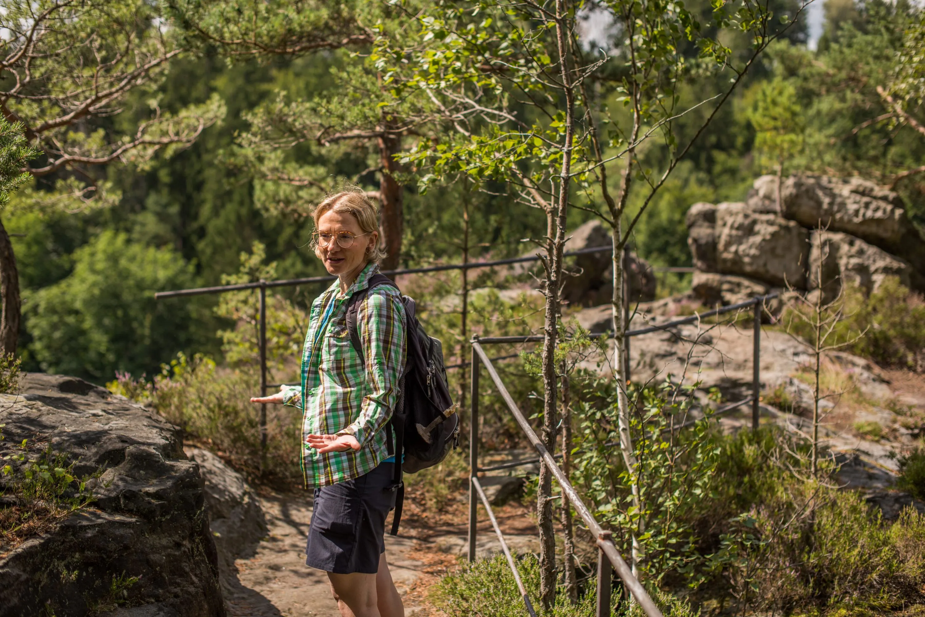 Eine Person mit einem Rucksack, die ein grĂŒnes Karohemd und Shorts trĂ€gt, lĂ€chelt, wĂ€hrend sie auf einem felsigen Waldweg mit MetallgelĂ€ndern wandert, umgeben von BĂ€umen und viel GrĂŒn.