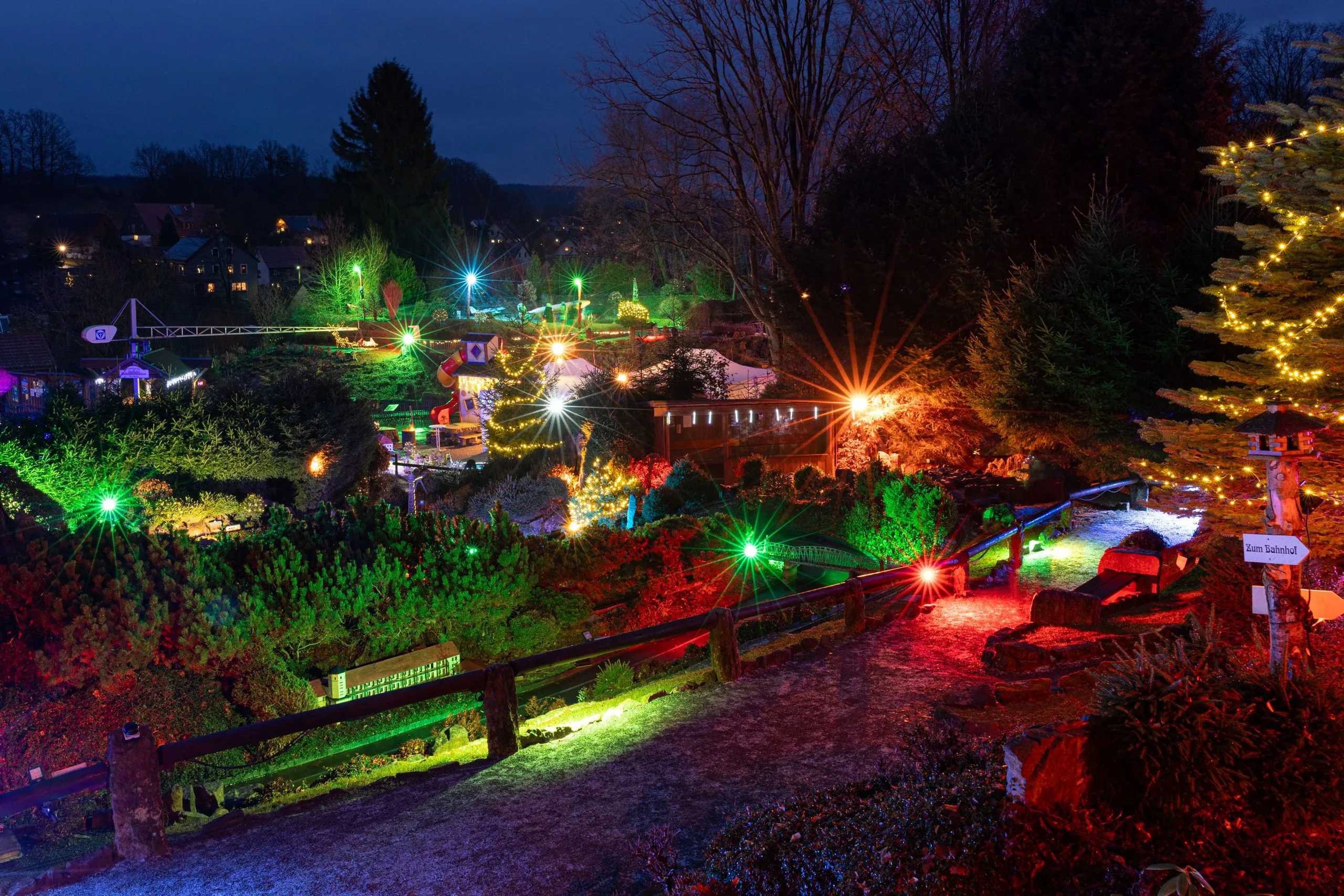 Eine nächtliche Gartenszene ist mit bunten Lichterketten geschmückt, die Bäume, Sträucher und Wege beleuchten. Rechts steht ein warm beleuchteter Weihnachtsbaum, und der dunkle Himmel sorgt für eine festliche Atmosphäre.