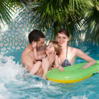Eine glückliche dreiköpfige Familie genießt einen Tag am Pool. Vater, Mutter und kleines Kind lächeln und planschen im Wasser. Das Kind sitzt auf einer grünen aufblasbaren Schwimmhilfe und wird von den Eltern gestützt. Üppige Palmwedel rahmen den Hintergrund ein.