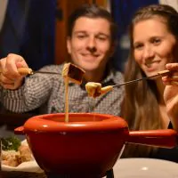 Ein Paar taucht bei einem Abendessen bei Kerzenschein Brot in einen roten Topf mit Käsefondue. Sie lächeln und sitzen eng beieinander, eine Weinflasche steht auf dem Tisch. Im Hintergrund sind Vorhänge und ein Fenster zu sehen, was eine intime, gemütliche Atmosphäre suggeriert.