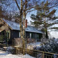 An einem verschneiten Tag steht eine rustikale Holzhütte mit schneebedecktem Dach umgeben von Bäumen. Links von der Hütte befindet sich eine umzäunte Terrasse mit Tisch und Stühlen. Um das Gebäude herum stehen hohe Kiefern, im Hintergrund ein strahlend blauer Himmel.
