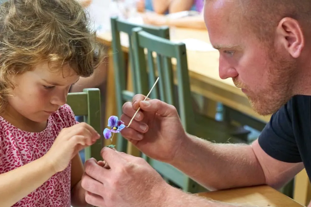 Ein Mann und ein junges Mädchen basteln an einem Tisch. Der Mann hält einen kleinen Stock und führt das Mädchen, das vorsichtig eine lila Blume auf ein kleines Objekt legt. Beide sind auf ihre Arbeit konzentriert. Im Hintergrund sind grüne Stühle und ein Holztisch zu sehen.