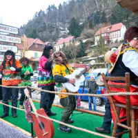 Auf einem lebhaften Festwagen sind mehrere Menschen in bunter Batikkleidung und Perücken zu sehen, die Gitarre spielen und tanzen. Eine weiß gekleidete Frau mit einer roten Schürze steht neben einer Holzbank. Im Hintergrund sind Häuser am Hang zu sehen und Wegweiser weisen auf verschiedene Städte hin.