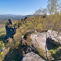 Ein Panoramablick auf eine von Bäumen umgebene Felsklippe in einem bewaldeten Tal bei Tag. Die Klippe zeichnet sich durch schroffe Felsformationen und Birken aus, im Hintergrund sind weite bewaldete Hügel und in der Ferne Berge unter einem klaren blauen Himmel zu sehen.