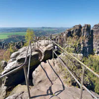 Panoramablick von einem Felsvorsprung mit Metallgeländer auf eine weite Landschaft mit üppigen grünen Feldern, fernen Hügeln und zerklüfteten Felsformationen. Der Himmel ist klar und Bäume säumen die umliegenden Felswände und unterstreichen die natürliche Schönheit der Gegend.