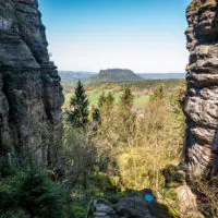 Eine malerische Aussicht auf das Elbsandsteingebirge zeigt eine schmale, felsige Schlucht, die ein in Sonnenlicht getauchtes Plateau in der Ferne umrahmt. Der Vordergrund zeigt üppiges Grün, mit einem gewundenen Pfad, der in die Schlucht hinunterführt, umgeben von hoch aufragenden Felsformationen und Bäumen.