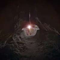 Ein Blick aus dem Inneren einer dunklen, felsigen Höhle mit einer schmalen Durchgangsöffnung. Sonnenlicht strömt durch die Öffnung, beleuchtet die Höhlenwände und erzeugt einen dramatischen Kontrast zwischen dem dunklen Inneren und dem hellen Licht draußen.