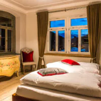 Ein gemütliches Schlafzimmer mit einem Doppelbett, das mit roten und grauen Kissen geschmückt ist. Eine Holzkommode mit Blumenmuster steht neben einem Sessel in der Nähe eines Fensters. Das Zimmer verfügt über elegante Vorhänge an den Fenstern, einen dekorativen Spiegel an der Wand und warme Beleuchtung.