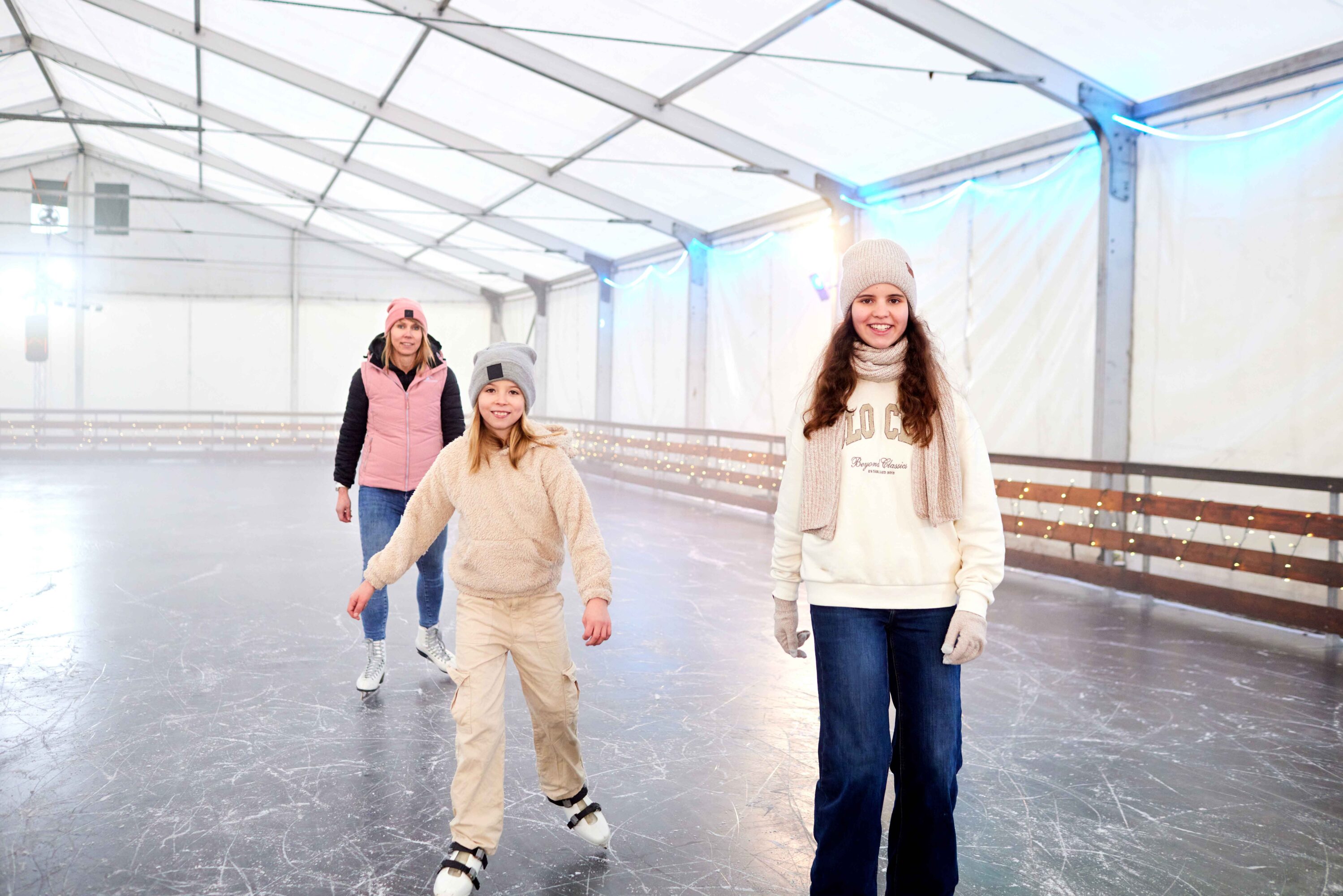 Drei in Winterkleidung und Mützen gekleidete Personen laufen gemeinsam auf einer überdachten Eisbahn Schlittschuh. Die Gruppe läuft lächelnd auf dem glatten Eis der Halle unter einem weißen Zelt mit blauen Lichtern auf die Kamera zu.