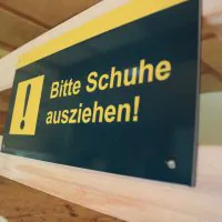 Eine Nahaufnahme eines rechteckigen Schildes mit gelbem Rand, das auf einer Holzoberfläche angebracht ist. Das Schild hat ein gelbes Ausrufezeichensymbol und lautet auf Deutsch „Bitte Schuhe ausziehen!“, was auf Englisch „Bitte ziehen Sie Ihre Schuhe aus!“ bedeutet.