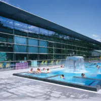An einem sonnigen Tag entspannen Menschen in einem Außenpool mit Springbrunnen. Der Pool grenzt an ein modernes Glasgebäude mit großen Fenstern. Im Hintergrund sind gelbe Liegestühle und grüne Bäume zu sehen, die zur ruhigen Atmosphäre beitragen.