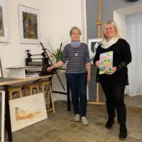 Zwei Frauen stehen in einem Kunstatelier, umgeben von gerahmten Kunstwerken. Die eine trägt ein gestreiftes Hemd und steht neben einem Tisch. Die andere, ganz in Schwarz gekleidet, lächelt, während sie ein buntes Buch in der Hand hält. Der Raum hat Fliesenboden und weiße Wände.