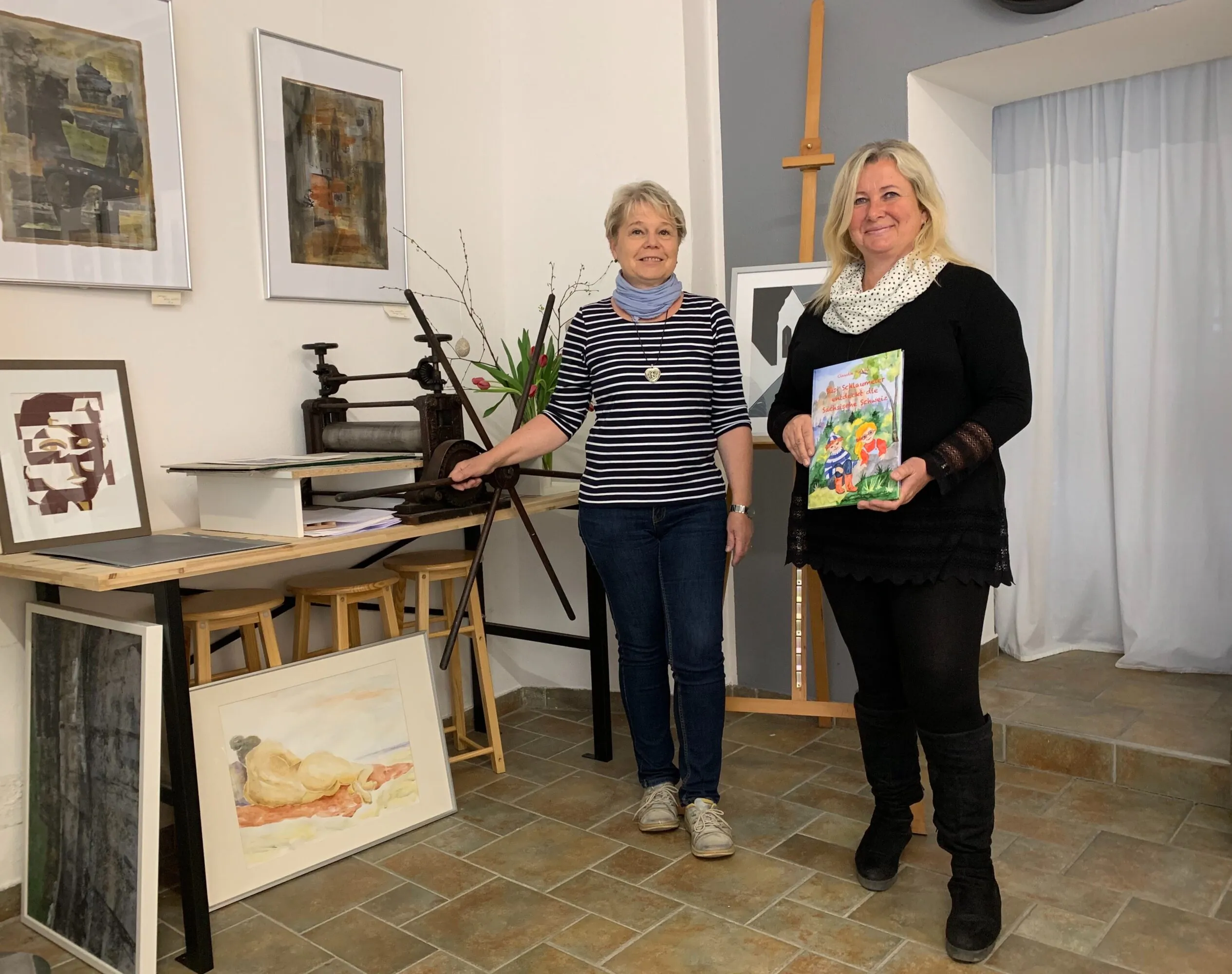 Zwei Frauen stehen in einem Kunstatelier, umgeben von gerahmten Kunstwerken. Die eine trägt ein gestreiftes Hemd und steht neben einem Tisch. Die andere, ganz in Schwarz gekleidet, lächelt, während sie ein buntes Buch in der Hand hält. Der Raum hat Fliesenboden und weiße Wände.