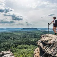 Eine Person mit einem Rucksack steht auf einem Felsvorsprung mit einem Metallgeländer und blickt unter einem teilweise bewölkten Himmel auf eine weitläufige Waldlandschaft.