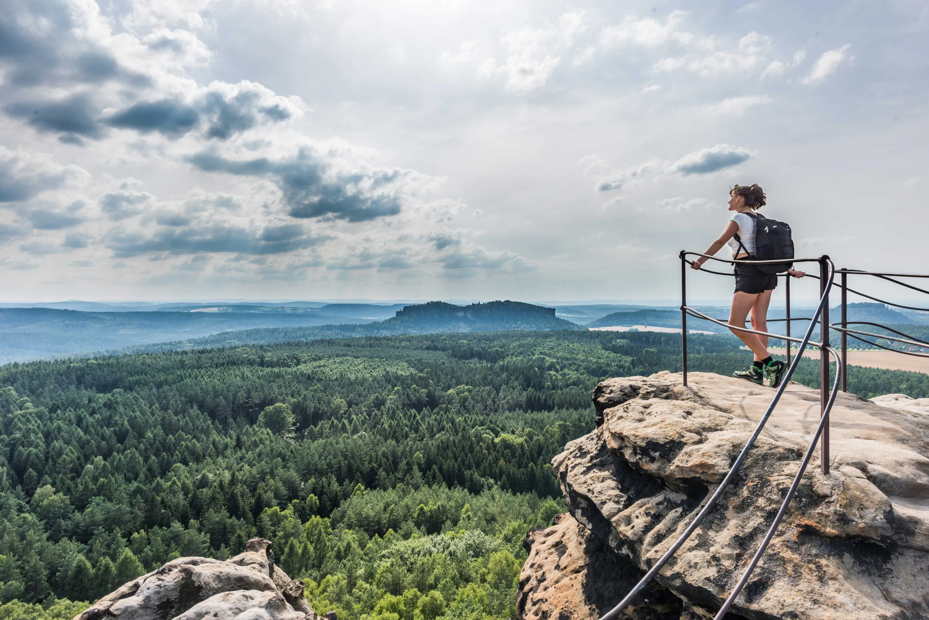 Eine Person mit einem Rucksack steht auf einem Felsvorsprung mit einem Metallgeländer und blickt unter einem teilweise bewölkten Himmel auf eine weitläufige Waldlandschaft.