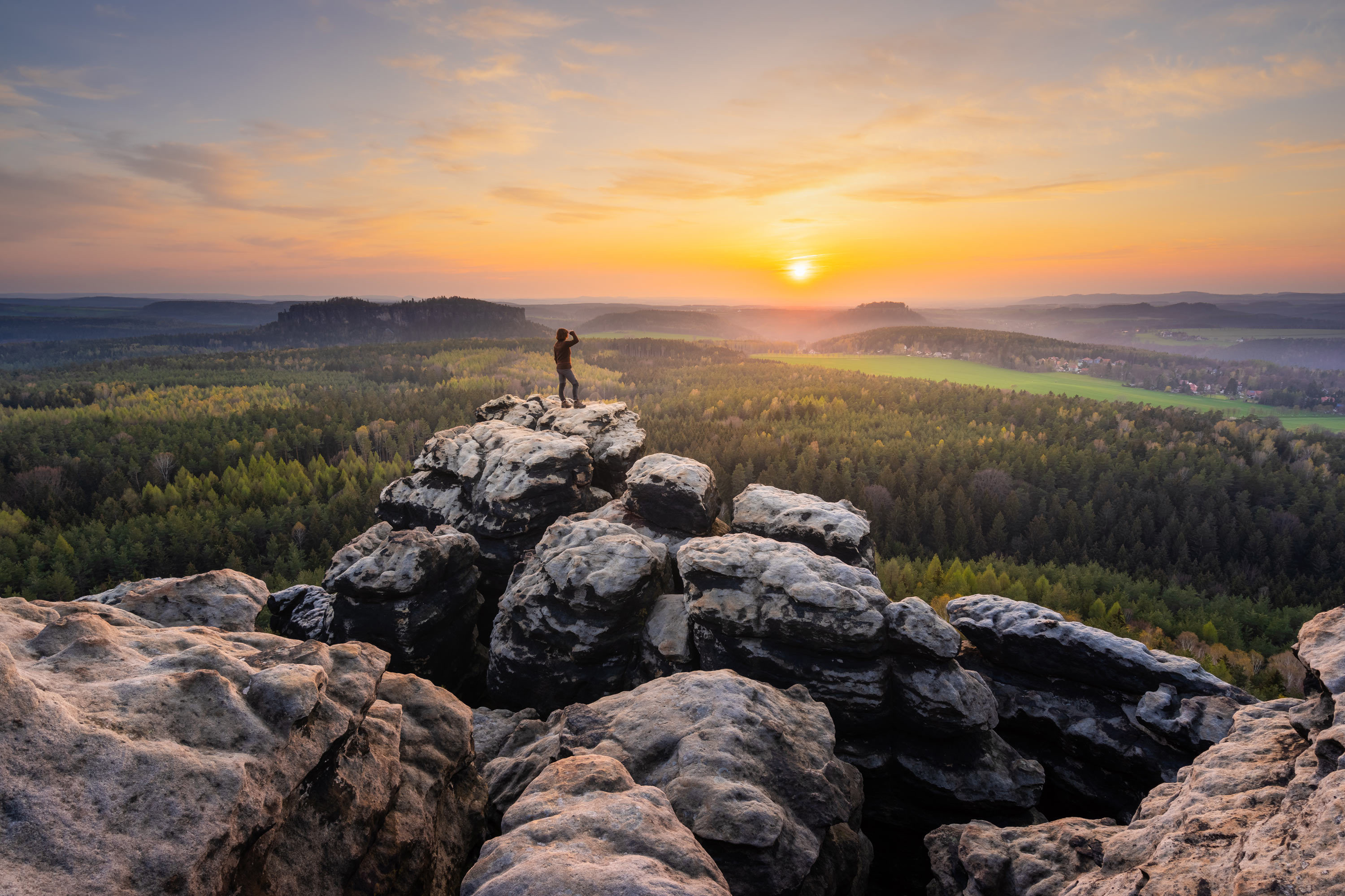 Eine Person steht auf zerklüftetem, felsigem Gelände, hält einen Hund an der Leine und blickt bei Sonnenaufgang auf einen riesigen, grünen Wald. Der Himmel ist in einer Mischung warmer Farben gehalten, als die Sonne über dem Horizont aufgeht und einen sanften, goldenen Schimmer über die Landschaft wirft.