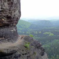Ein schmaler, steiniger Pfad schlängelt sich am Rand einer steilen Klippe entlang und bietet einen malerischen Blick auf ein üppiges, grünes Tal und sanfte Hügel unter einem bewölkten Himmel. Die umliegende Landschaft ist dicht bewaldet, wobei unterschiedliche Grüntöne auf die vielfältige Vegetation darunter hinweisen.