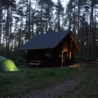 Eine kleine Holzhütte mit eingeschaltetem Licht steht mitten in einem Wald mit hohen Bäumen. In der Nähe steht ein grünes Zelt und um die Hütte herum stehen Picknicktische. Der Himmel verdunkelt sich und kündigt die nahende Dämmerung an.