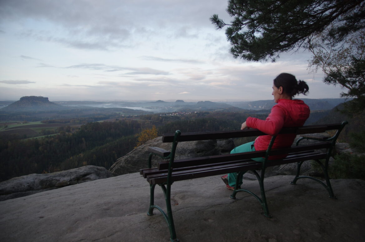 Eine Person in einer roten Jacke sitzt auf einer Bank und blickt vom Rand einer Klippe auf eine malerische Landschaft. Die Aussicht umfasst sanfte Hügel, einen Wald und ein entferntes Plateau unter einem bewölkten Himmel. Ein Ast umrahmt teilweise die obere rechte Ecke des Bildes.