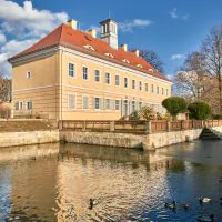 Ein großes beiges Gebäude mit rotem Dach steht neben einem ruhigen Teich mit Enten. Das Gebäude ist von einer Steinterrasse umgeben und die Bäume sind jetzt kahl, was auf Winter hindeutet. Der Himmel ist klar und blau mit ein paar vereinzelten Wolken.