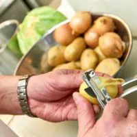 Eine Person schält in einer Küche eine Kartoffel mit einem Schäler. Im Hintergrund stehen Schüsseln mit ungeschälten Kartoffeln, Zwiebeln und einem Kohlkopf. Die Person trägt eine Armbanduhr aus Edelstahl.
