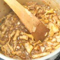 Ein Topf mit einer Mischung aus geschnittenem Kohl und Zwiebeln wird mit einem Holzspatel umgerührt. Die Zutaten kochen in einer braunen Soße und scheinen teilweise gekocht zu sein. Ein Teil der metallischen Oberfläche des Topfes ist sichtbar, ebenso wie etwas Arbeitsfläche.