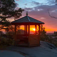 Auf einem felsigen Hügel, umgeben von Bäumen, steht ein Pavillon aus Holz, von dem aus man einen herrlichen Sonnenuntergang mit bunten Wolken am Himmel bewundern kann.