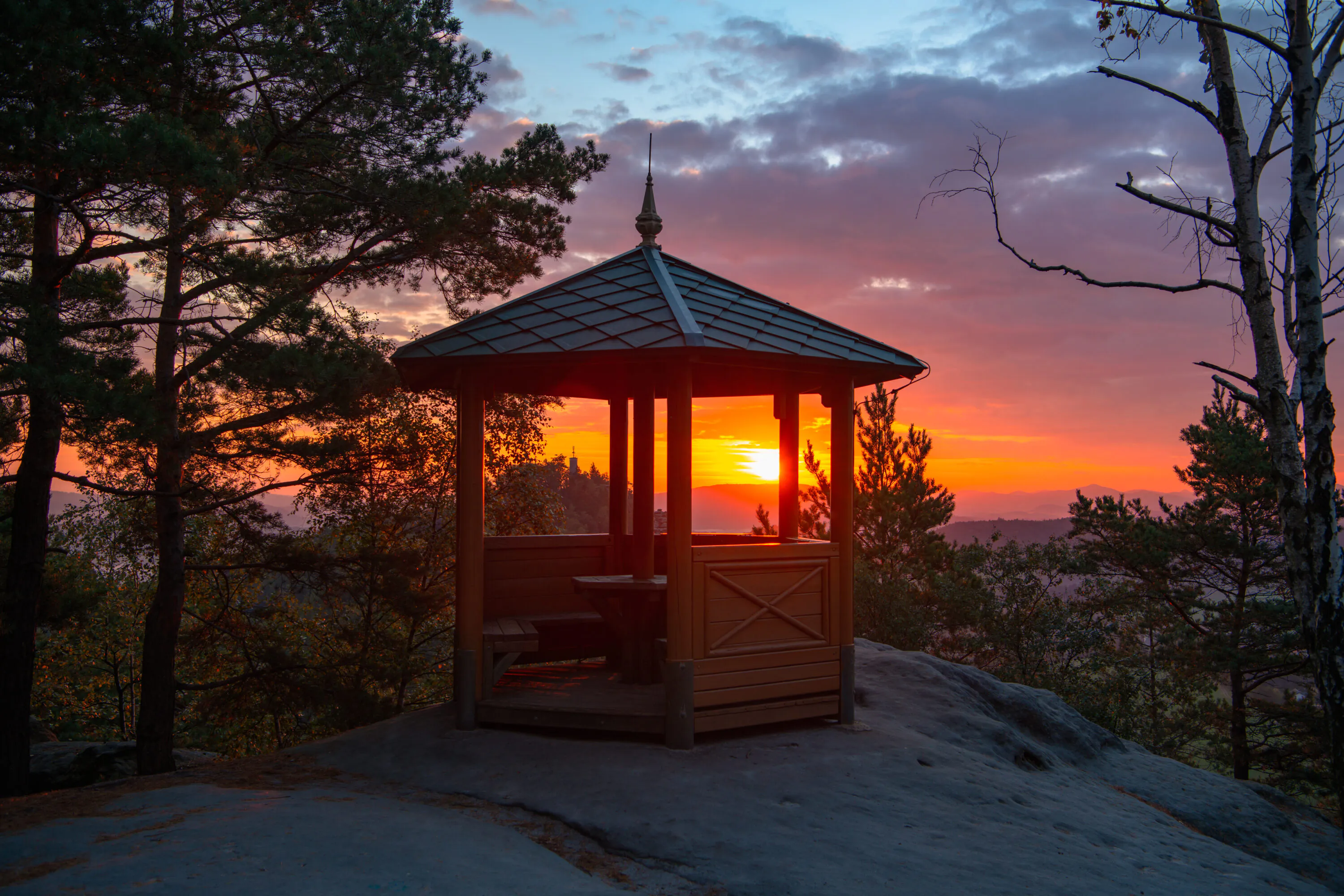 Auf einem felsigen Hügel, umgeben von Bäumen, steht ein Pavillon aus Holz, von dem aus man einen herrlichen Sonnenuntergang mit bunten Wolken am Himmel bewundern kann.