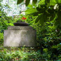 Ein Grabstein mit der Inschrift „Robert Sterl“, umgeben von üppigem Grün und Bäumen. Auf dem Grabstein steht ein runder Blumenkübel mit roten Blumen.