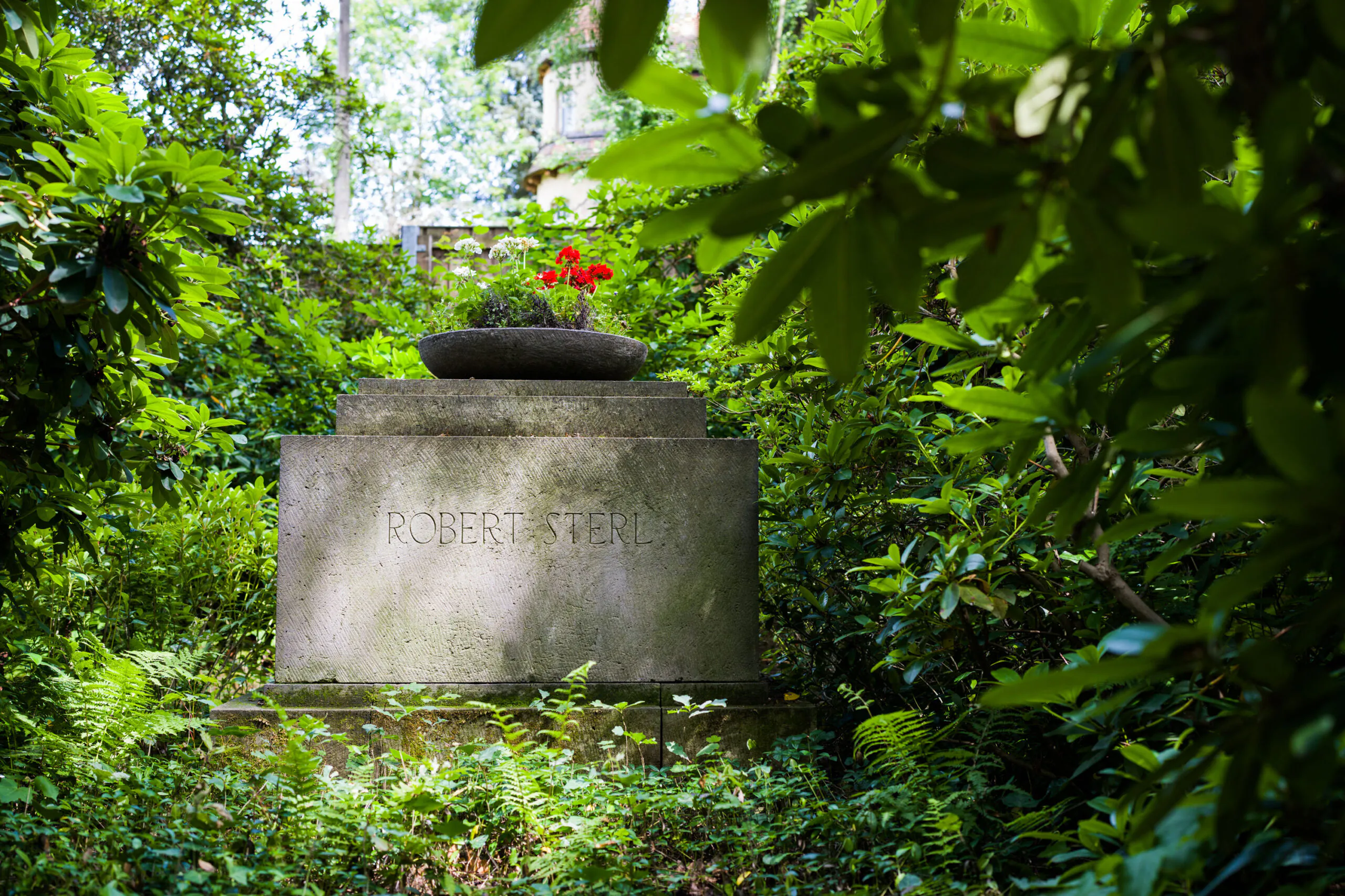 Ein Grabstein mit der Inschrift „Robert Sterl“, umgeben von üppigem Grün und Bäumen. Auf dem Grabstein steht ein runder Blumenkübel mit roten Blumen.