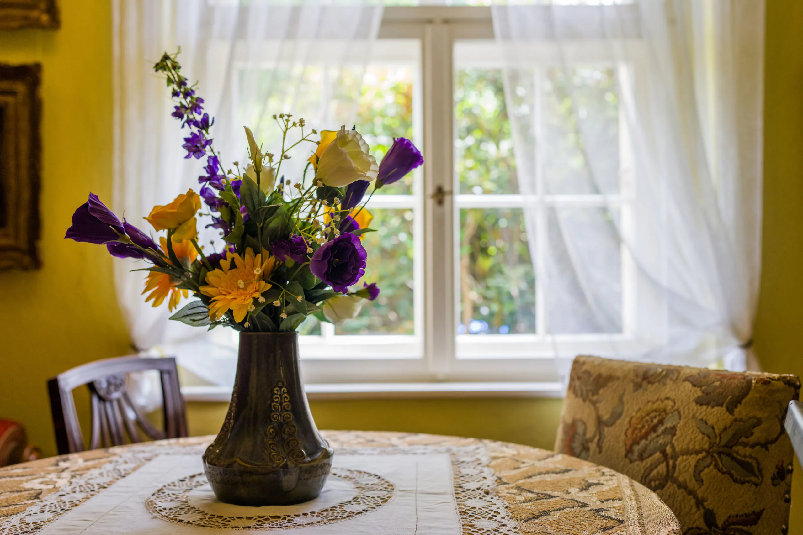 Auf einem runden Tisch, der mit einer Spitzentischdecke bedeckt ist, steht eine Vase mit bunten Blumen vor einem sonnendurchfluteten Fenster mit weißen, durchsichtigen Vorhängen.