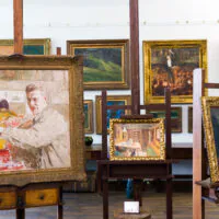In einer Kunstgalerie auf Staffeleien ausgestellte Gemälde, darunter ein Porträt eines Mannes im Vordergrund, umgeben von verschiedenen gerahmten Kunstwerken an den Wänden eines gut beleuchteten Raums.