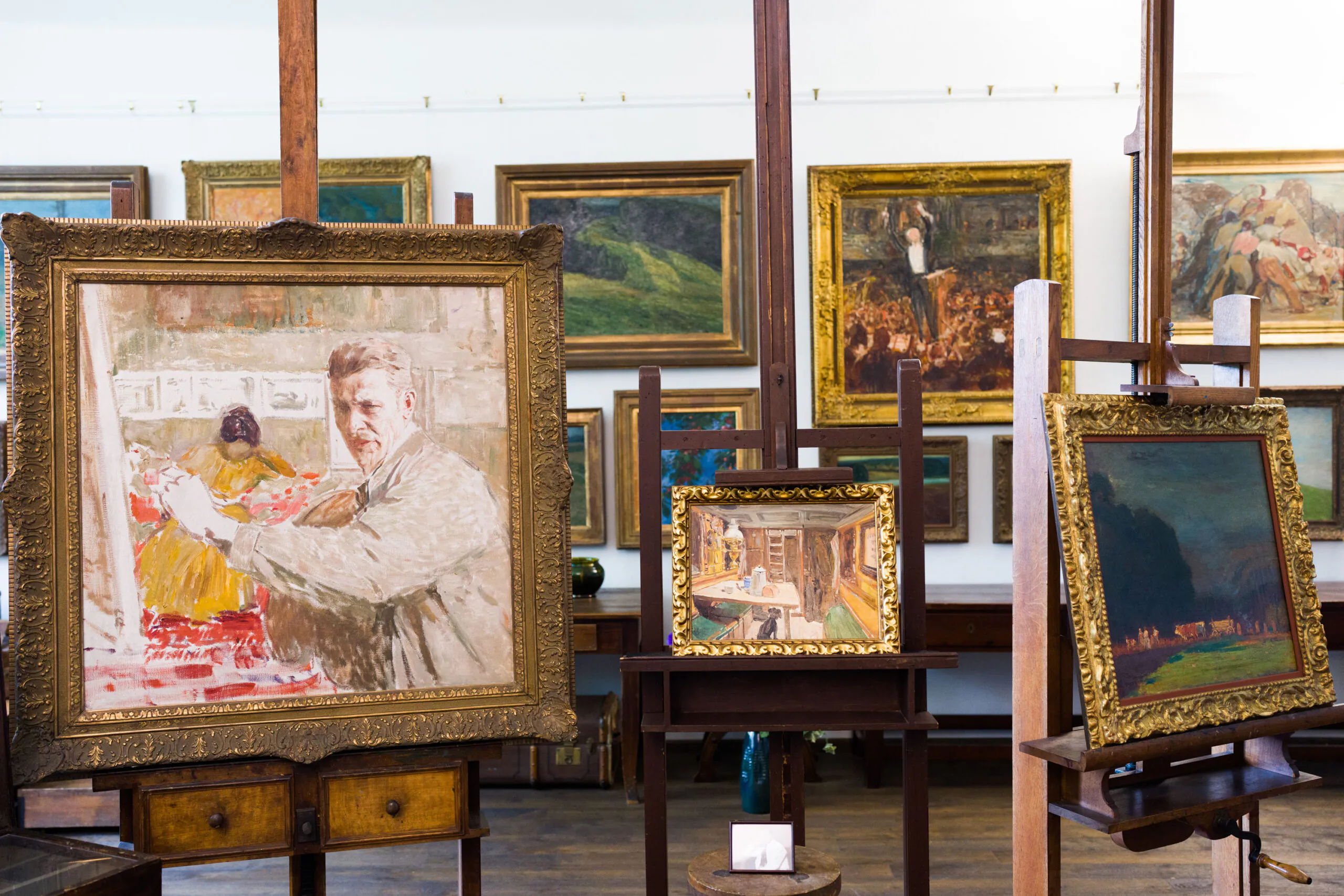 In einer Kunstgalerie auf Staffeleien ausgestellte Gemälde, darunter ein Porträt eines Mannes im Vordergrund, umgeben von verschiedenen gerahmten Kunstwerken an den Wänden eines gut beleuchteten Raums.