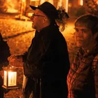 Eine Gruppe von Menschen steht nachts im Freien, warm angezogen. Eine Person in der Mitte hält eine brennende Laterne in der Hand und trägt einen Hut und einen Umhang. Die Szene wird von einem sanften, orangefarbenen Schein erhellt, auf dem Boden sind abgefallene Blätter zu sehen.
