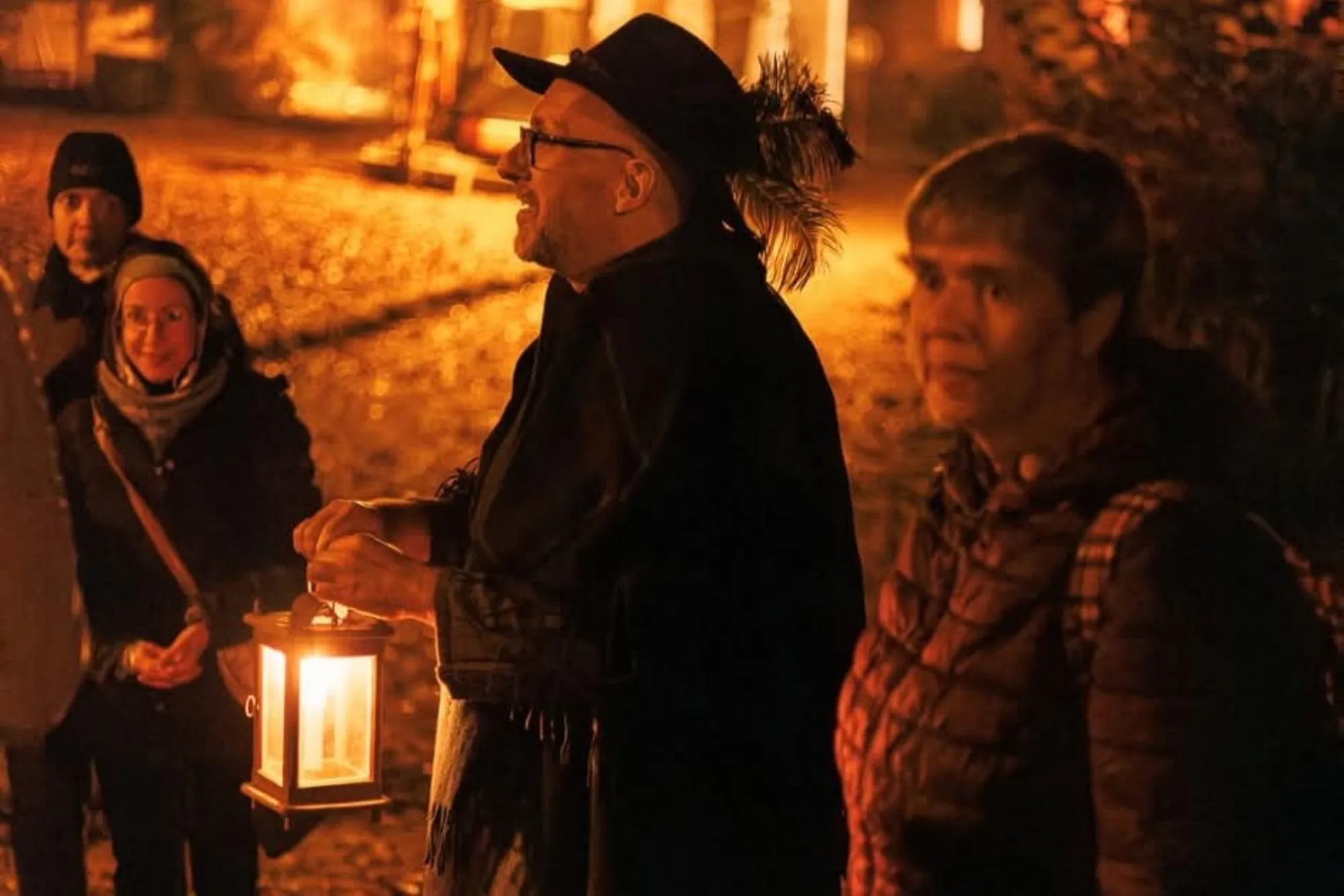 Eine Gruppe von Menschen steht nachts im Freien, warm angezogen. Eine Person in der Mitte hält eine brennende Laterne in der Hand und trägt einen Hut und einen Umhang. Die Szene wird von einem sanften, orangefarbenen Schein erhellt, auf dem Boden sind abgefallene Blätter zu sehen.