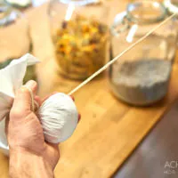 Die Hand einer Person hält ein Stoffbündel mit einer Schnur, das möglicherweise Kräuter oder Gewürze enthält. Im Hintergrund stehen drei offene Gläser mit verschiedenen getrockneten Pflanzen oder Blumen auf einer Holzoberfläche. Das Foto hat ein Wasserzeichen mit der Aufschrift „ACHIMEURER HDR PHOTOGRAPHY“.