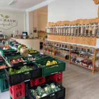 Ein Zero-Waste-Lebensmittelladen mit frischen Produkten in grünen Kisten, Abfüllbehältern an einer Holzwand und Regalen mit Gläsern und Konserven. Der Laden wirkt sauber und aufgeräumt.