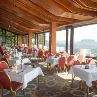 Ein geräumiges Restaurant mit rot-goldenen Stühlen, weißen Tischdecken, großen Fenstern, die einen malerischen Blick auf die fernen Berge bieten, und Holzbalken an der Decke. Der Raum ist von natürlichem Licht durchflutet.