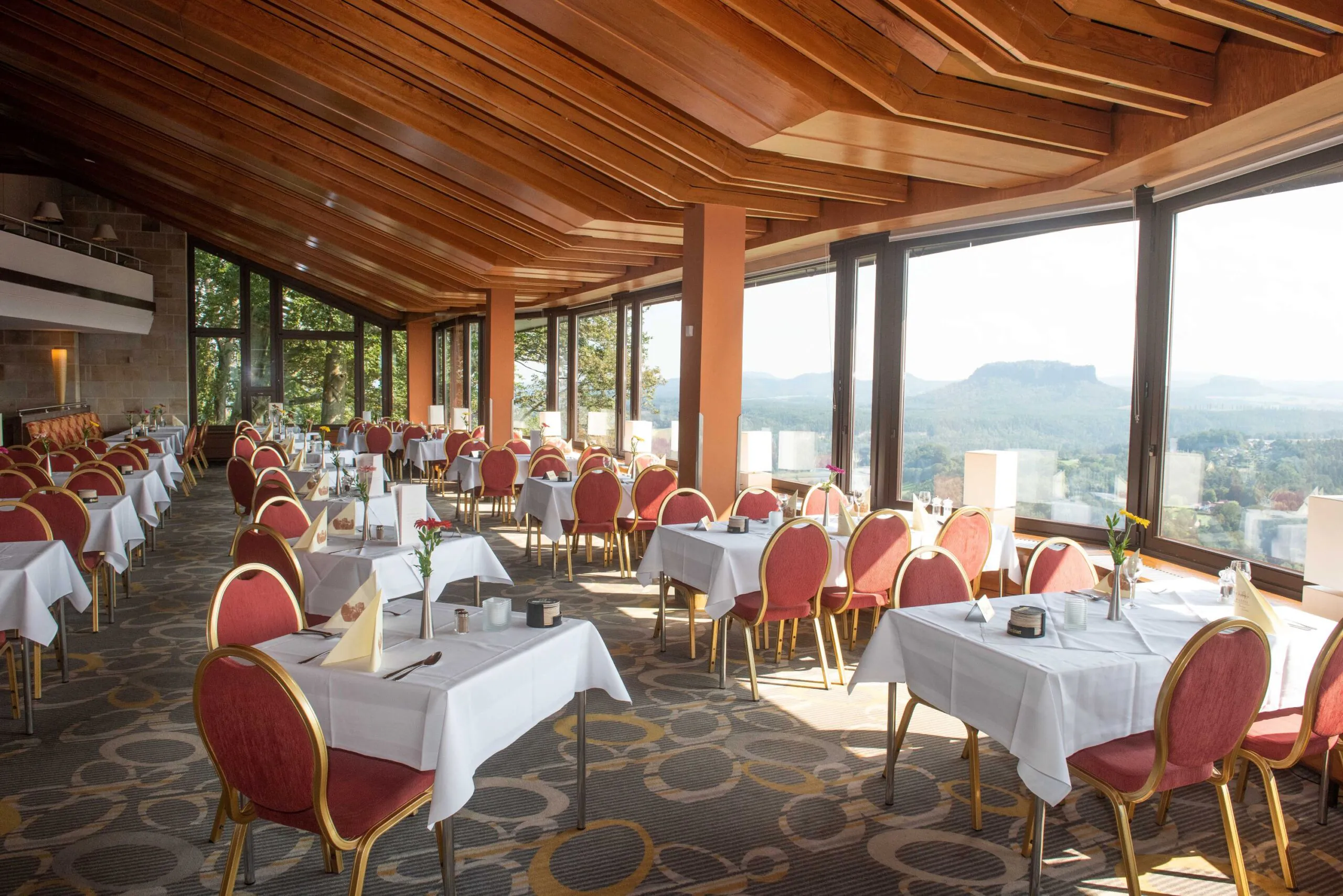 Ein geräumiges Restaurant mit rot-goldenen Stühlen, weißen Tischdecken, großen Fenstern, die einen malerischen Blick auf die fernen Berge bieten, und Holzbalken an der Decke. Der Raum ist von natürlichem Licht durchflutet.