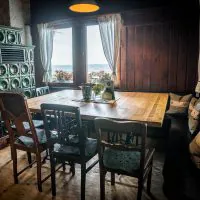 Ein gemütlicher Essbereich mit einem Holztisch, umgeben von verschiedenen Holzstühlen. Die Deckenleuchte wirft einen warmen Schein über den rustikal eingerichteten Raum. Ein grüner Keramikofen steht in der Ecke neben Fenstern mit weißen Vorhängen und bietet einen malerischen Blick nach draußen.