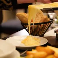 Geschmolzener Käse wird von einem Raclette-Grill auf einen weißen Teller geschabt, im Hintergrund sind Brot und Käse zu sehen.