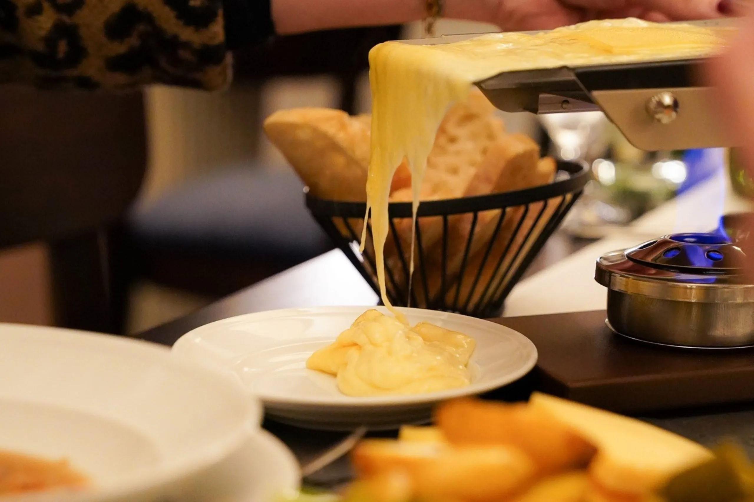 Geschmolzener Käse wird von einem Raclette-Grill auf einen weißen Teller geschabt, im Hintergrund sind Brot und Käse zu sehen.