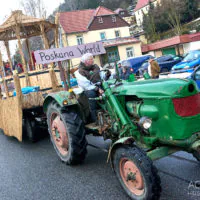 Ein Mann fährt einen grünen Traktor, der einen Festwagen mit Strohdach und einem Schild mit der Aufschrift „Poskana World“ zieht, bei einer Straßenparade. Schneeflecken sind sichtbar und Menschen schauen vom Bürgersteig aus zu.