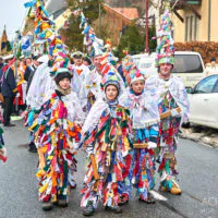 Eine Gruppe von Menschen in farbenfrohen, mit Bändern geschmückten Trachten und spitzen Hüten läuft in einer Parade durch eine Straße. Sie sind von Passanten und geparkten Autos umgeben. Die Gebäude im Hintergrund lassen eine Kleinstadtkulisse vermuten.