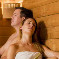 Zwei Menschen sitzen mit geschlossenen Augen und entspanntem Gesichtsausdruck in einer Holzsauna. Die Frau im Vordergrund ist in ein Handtuch gehüllt, während der Mann hinter ihr mit freiem Oberkörper dasteht. Warmes, sanftes Licht verstärkt die wohltuende Atmosphäre.