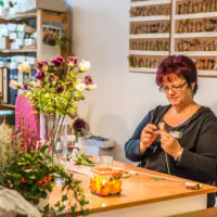 Eine Frau sitzt in einem hell erleuchteten Raum an einem Tisch und bastelt mit Blumen. Um sie herum stehen Vasen mit verschiedenen Blumen und Bastelutensilien. Dahinter sieht man Regale voller kleiner Blumentöpfe. Sie trägt eine Brille und hat kurze rote Haare und ist auf ihre Arbeit konzentriert.