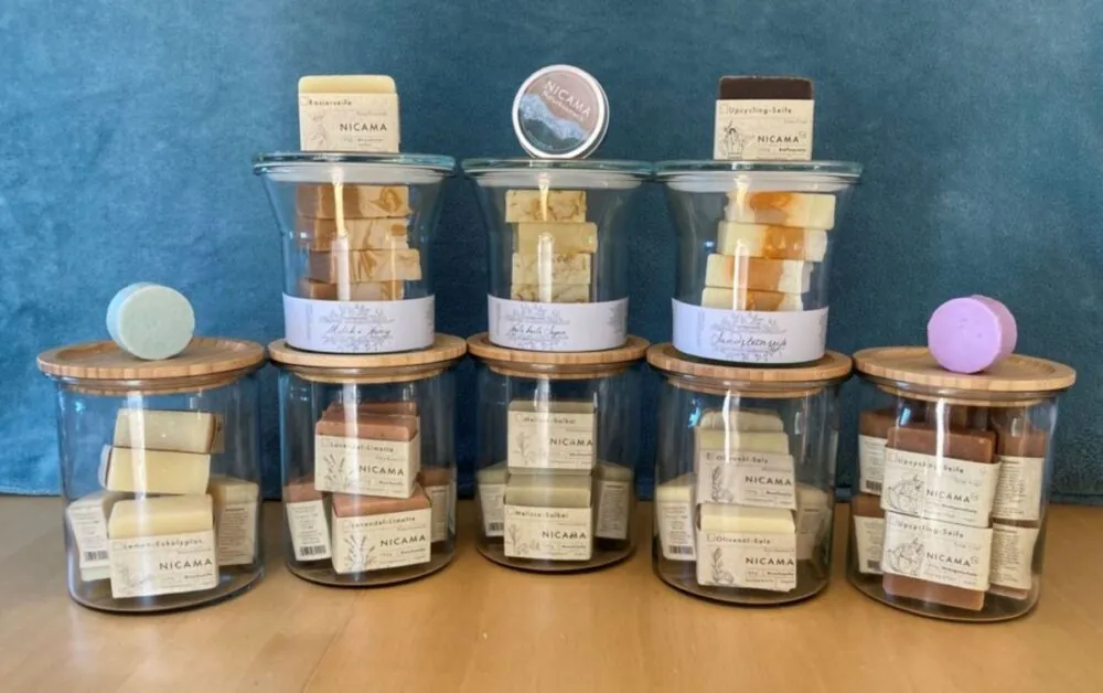 Neun Gläser mit Holzdeckeln, die jeweils mit handgemachten Seifenstücken in verschiedenen Farben gefüllt sind. Auf oder neben den Gläsern liegen zwei runde Seifenstücke und eine kleine Dose. Den Hintergrund bildet ein blauer Stoff.