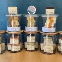 Neun Gläser mit Holzdeckeln, die jeweils mit handgemachten Seifenstücken in verschiedenen Farben gefüllt sind. Auf oder neben den Gläsern liegen zwei runde Seifenstücke und eine kleine Dose. Den Hintergrund bildet ein blauer Stoff.